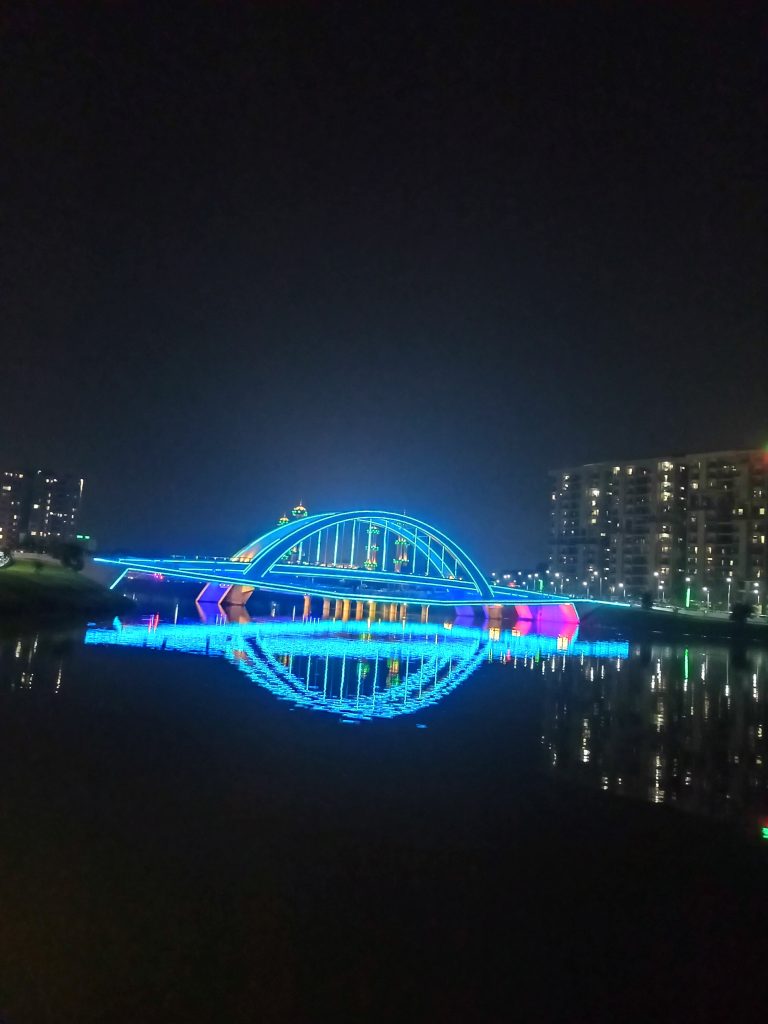 Neon-colored lights on a bridge reflected in water at night.