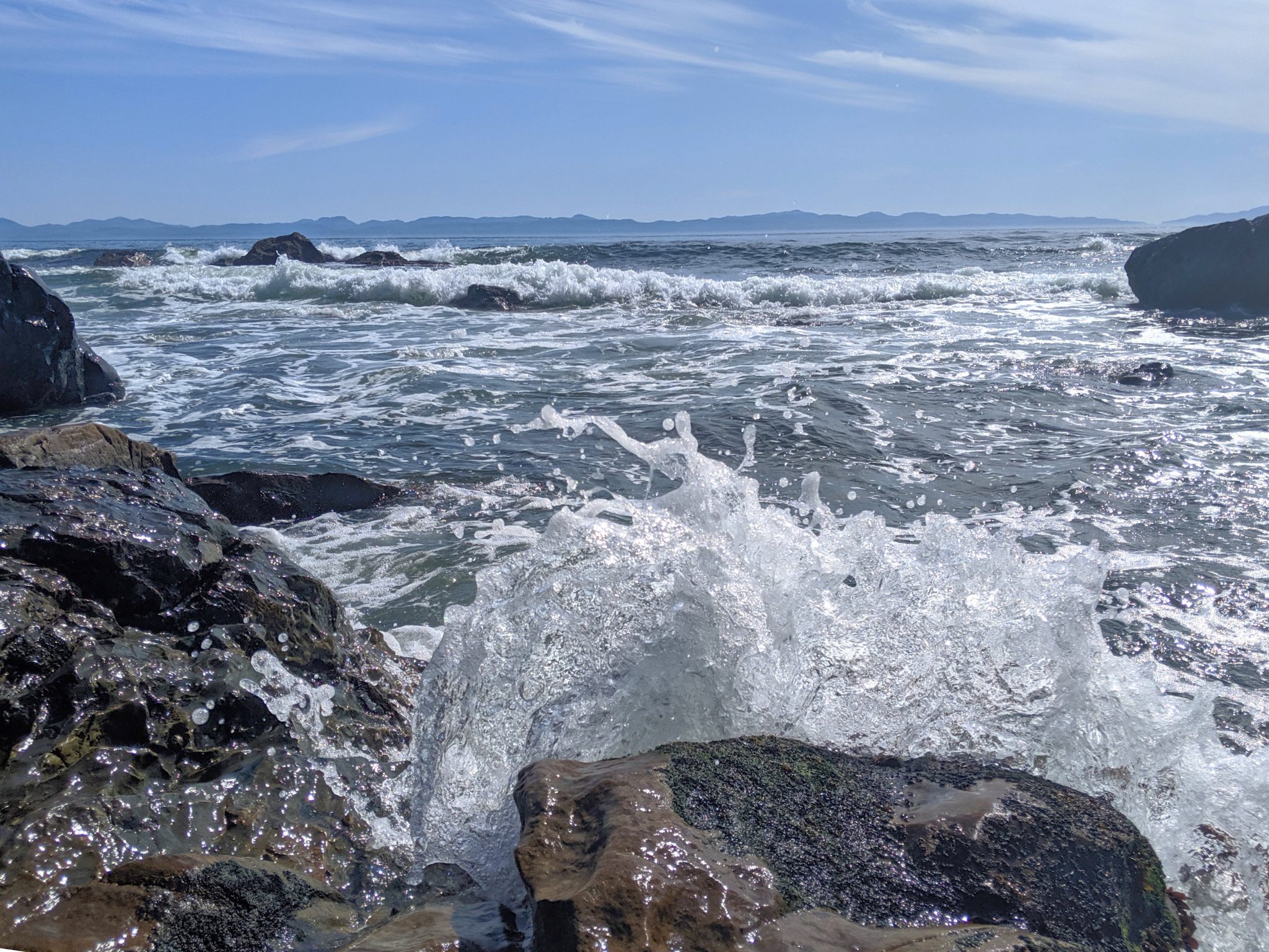 White ocean waves break over dark grey rocks along a coastline.