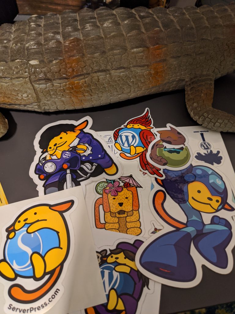 A pile of WordPress and WorCamp Wappu stickers on a table in front of a plastic alligator