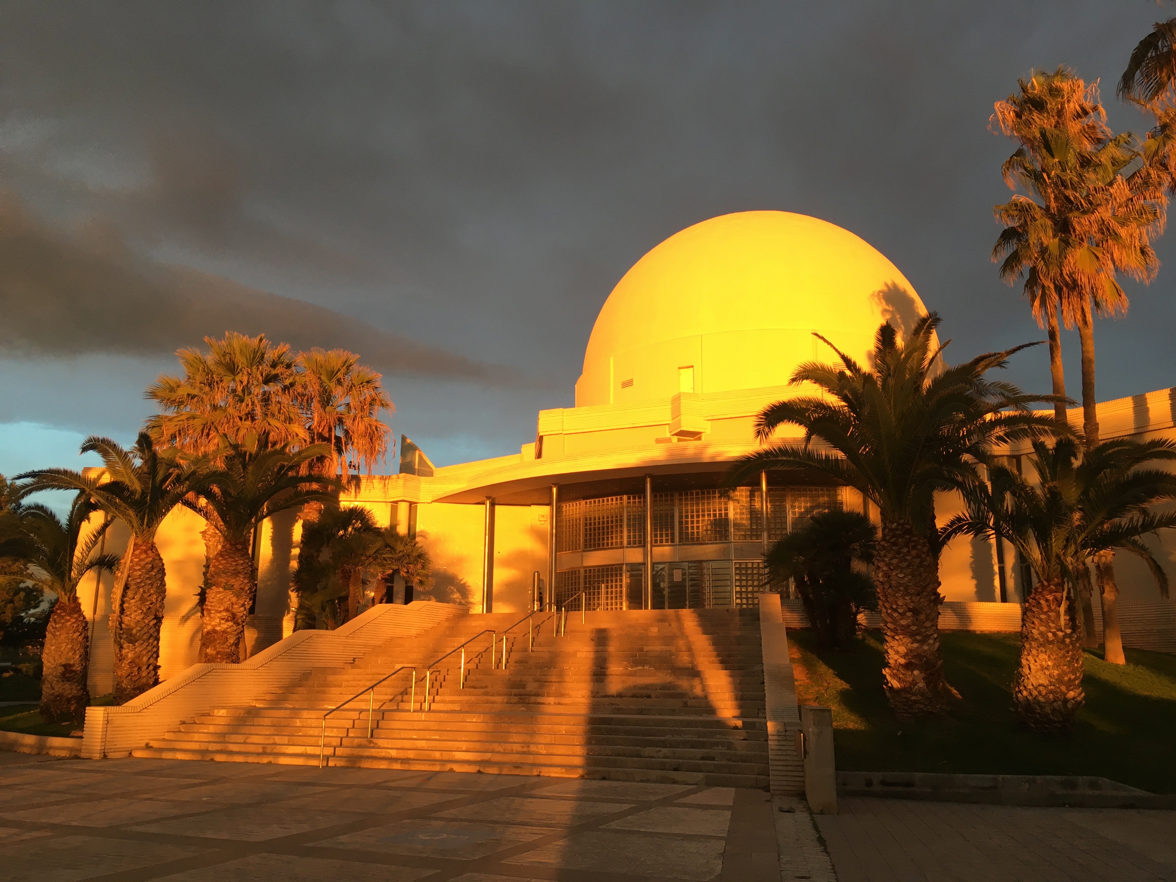 A large dome-shaped building surrounded by palm trees, bathed in warm sunlight. The entrance features wide steps with railings leading up to the building, and a dramatic sky with dark clouds adds depth to the scene.