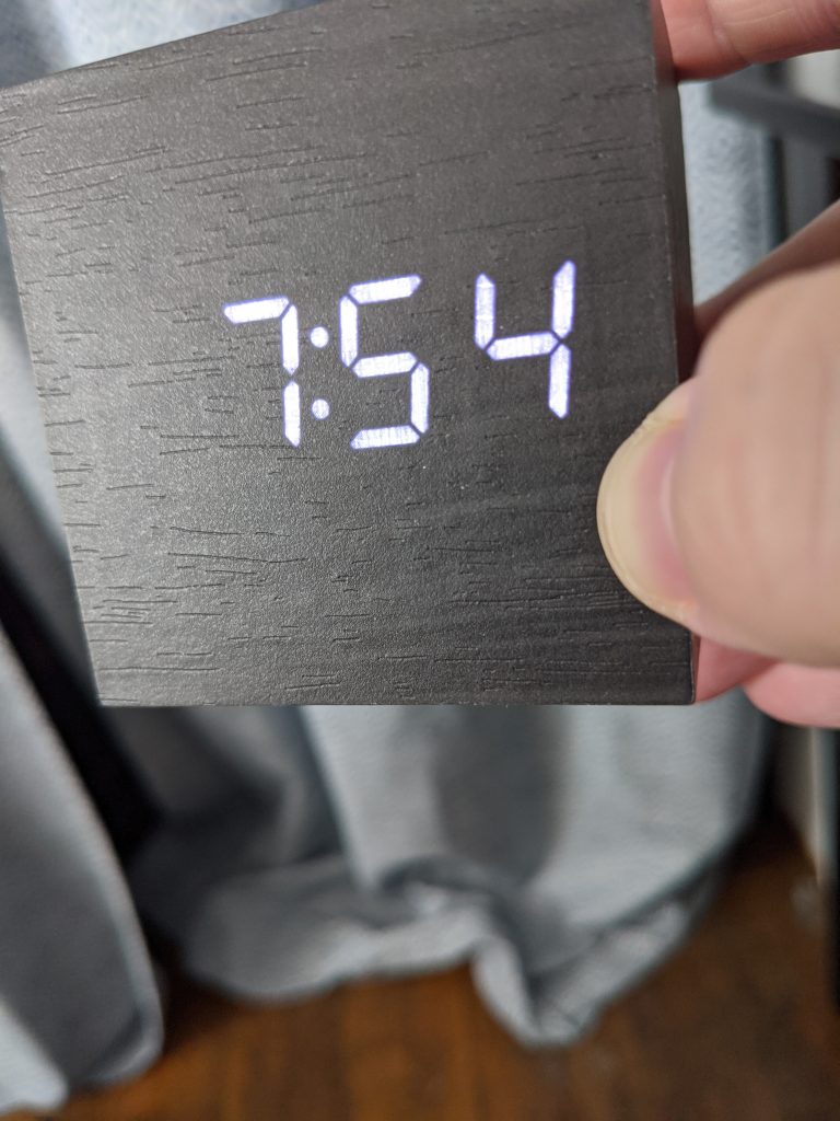 A square digital clock, held in someone’s hand. It is 7:54