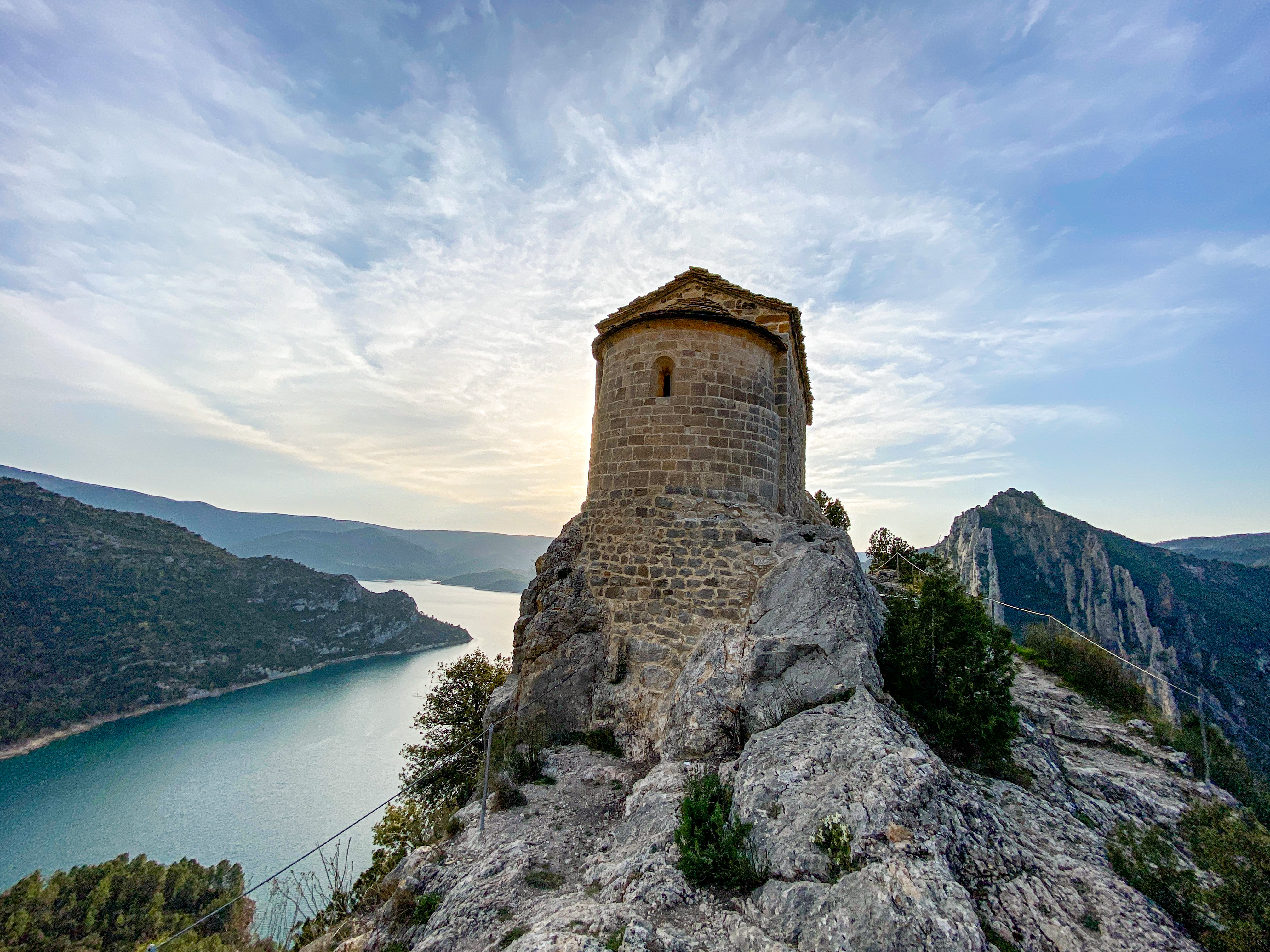 A small, round stone building perched on a rocky cliff overlooking a serene body of water, surrounded by mountains under a partly cloudy sky.