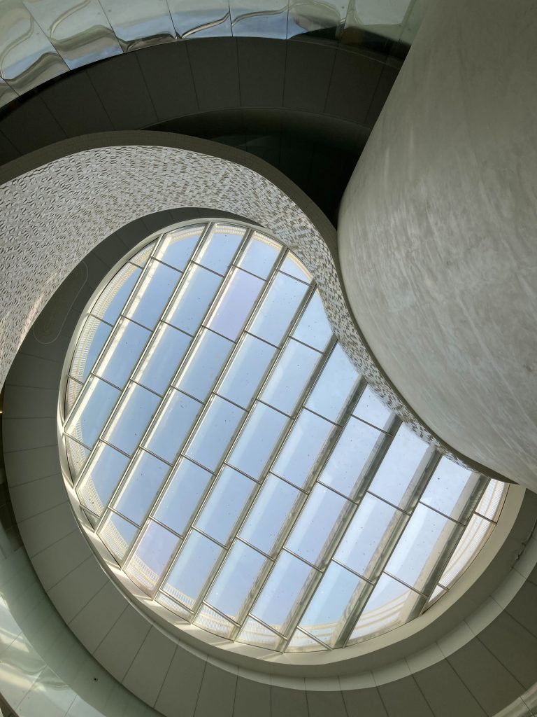 Curved architectural ceiling with a large, segmented glass skylight allowing natural light to enter. The surfaces surrounding the skylight appear smooth and feature modern design elements.