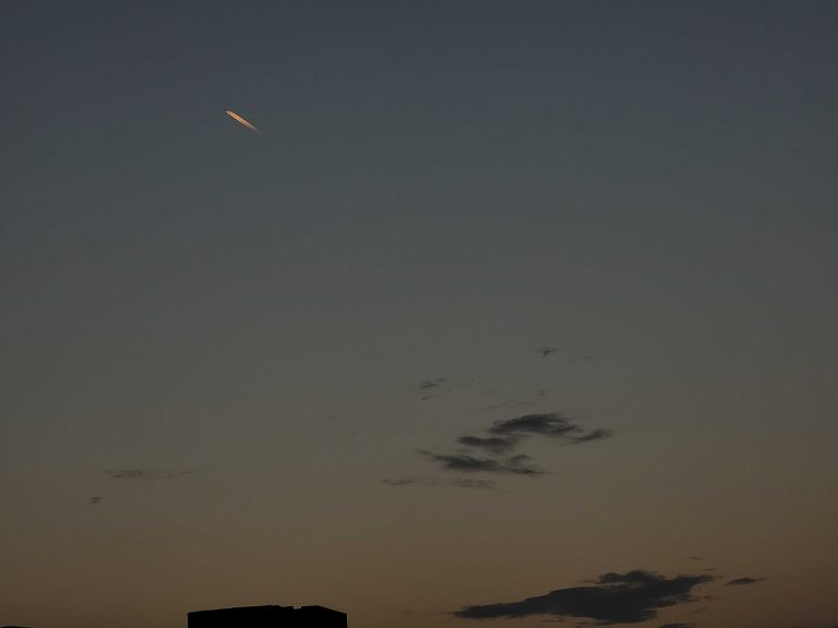 A darkening sky at dusk with a faintly illuminated object visible in the upper left, and silhouettes of clouds and a building in the lower portion.