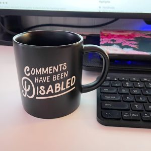 A mug with the text, “Comments Have Been Disabled” on it, next to a keyboard and a laptop on a cooling mat behind it
