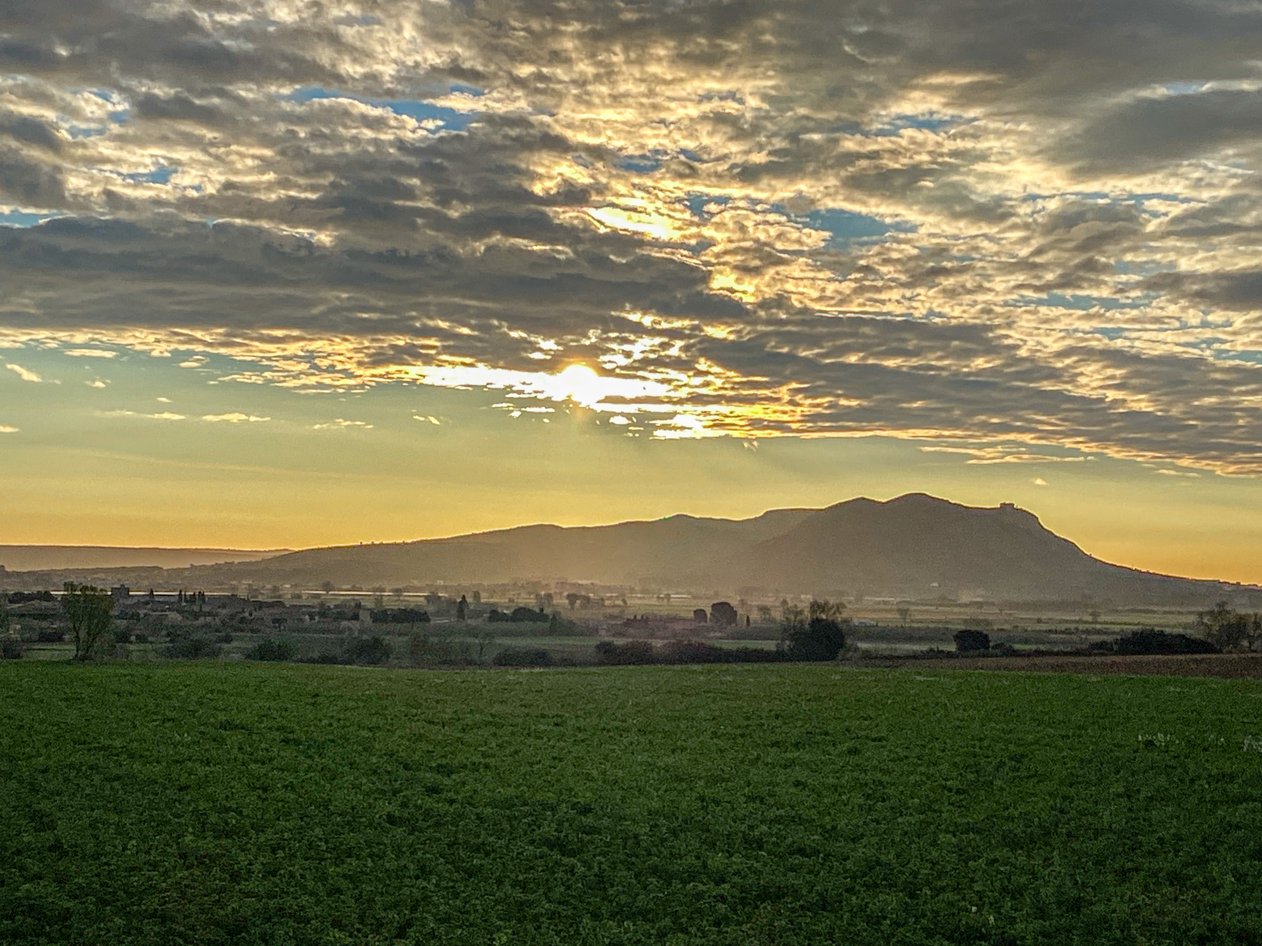 A scenic landscape featuring a lush green field in the foreground, with a distant mountain range under a dramatic sky. The sun is partially obscured by clouds, casting a warm, golden light over the scene.