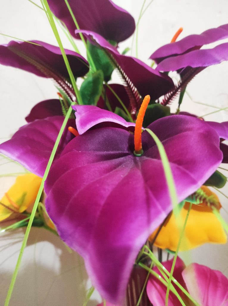 A close-up view of vibrant artificial flowers, predominantly in shades of purple with green stems and thin leaves. An orange stamen is visible protruding from the center of the main flower.