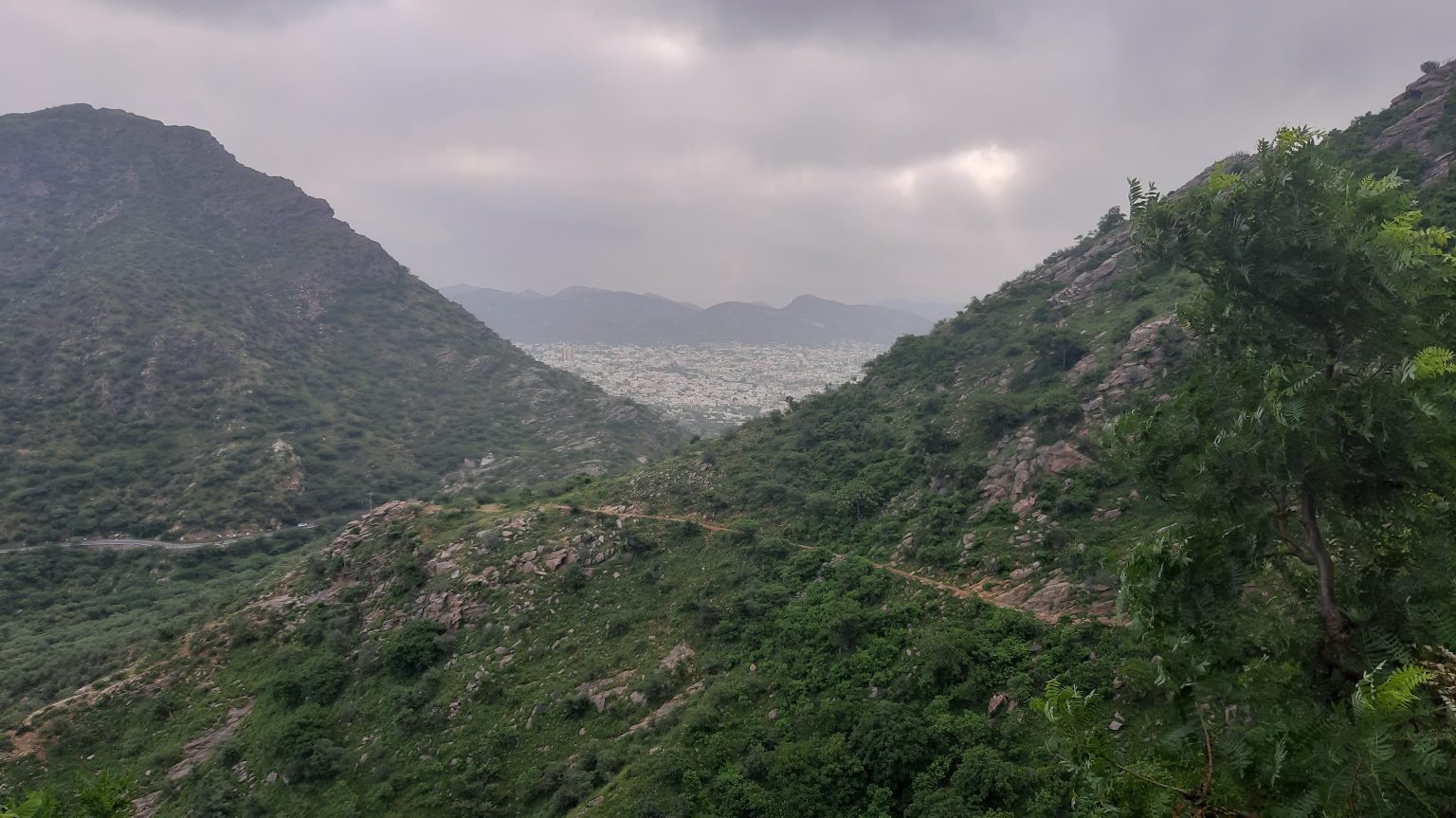 A lush green valley with rocky hills covered in vegetation, leading to a distant view of a cityscape under a cloudy sky.