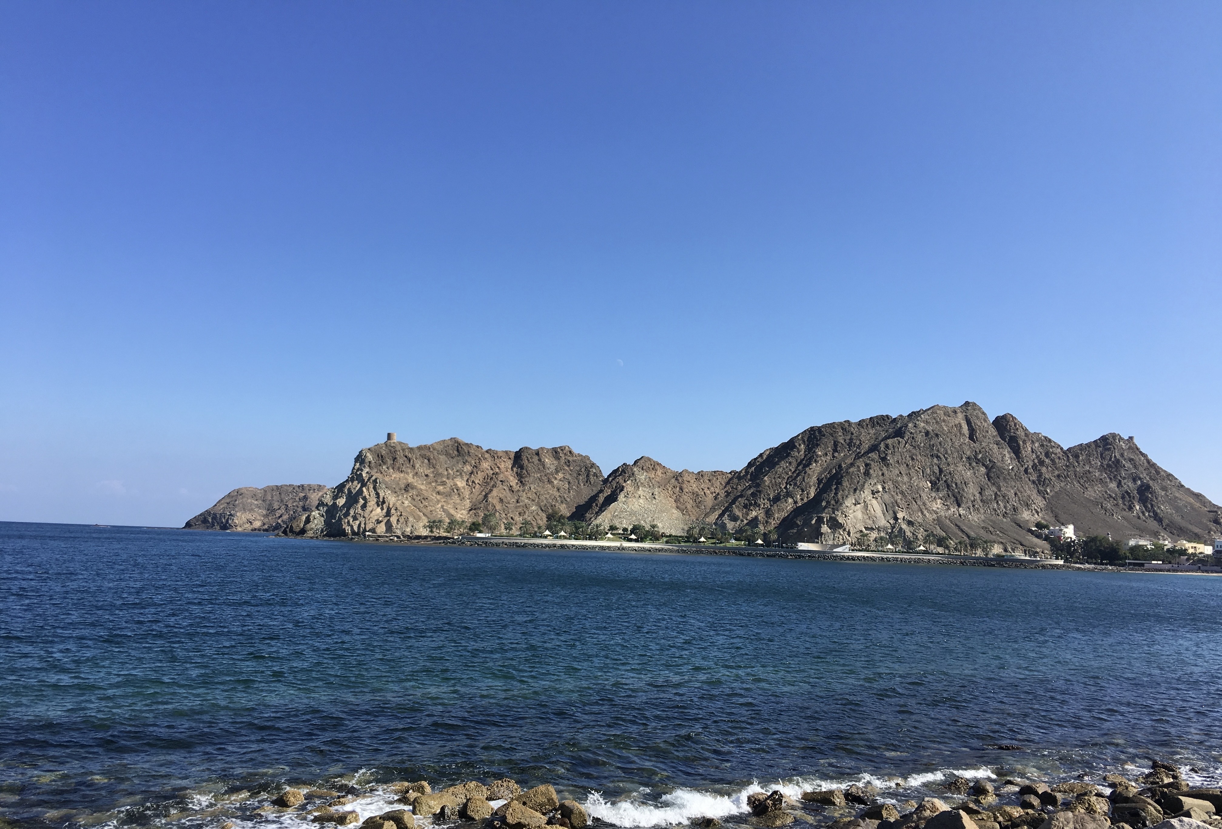 A scenic coastal view featuring a clear blue sky and calm turquoise waters. In the background, rugged mountains rise sharply from the shore, with one peak showing a small tower atop it. The foreground includes a rocky shoreline with clear water lapping against the stones, creating a serene and picturesque natural landscape.