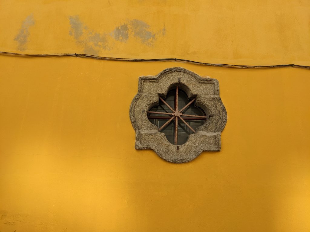 Yellow wall with porthole style window and wires with some dappled shadow and faded paint.
