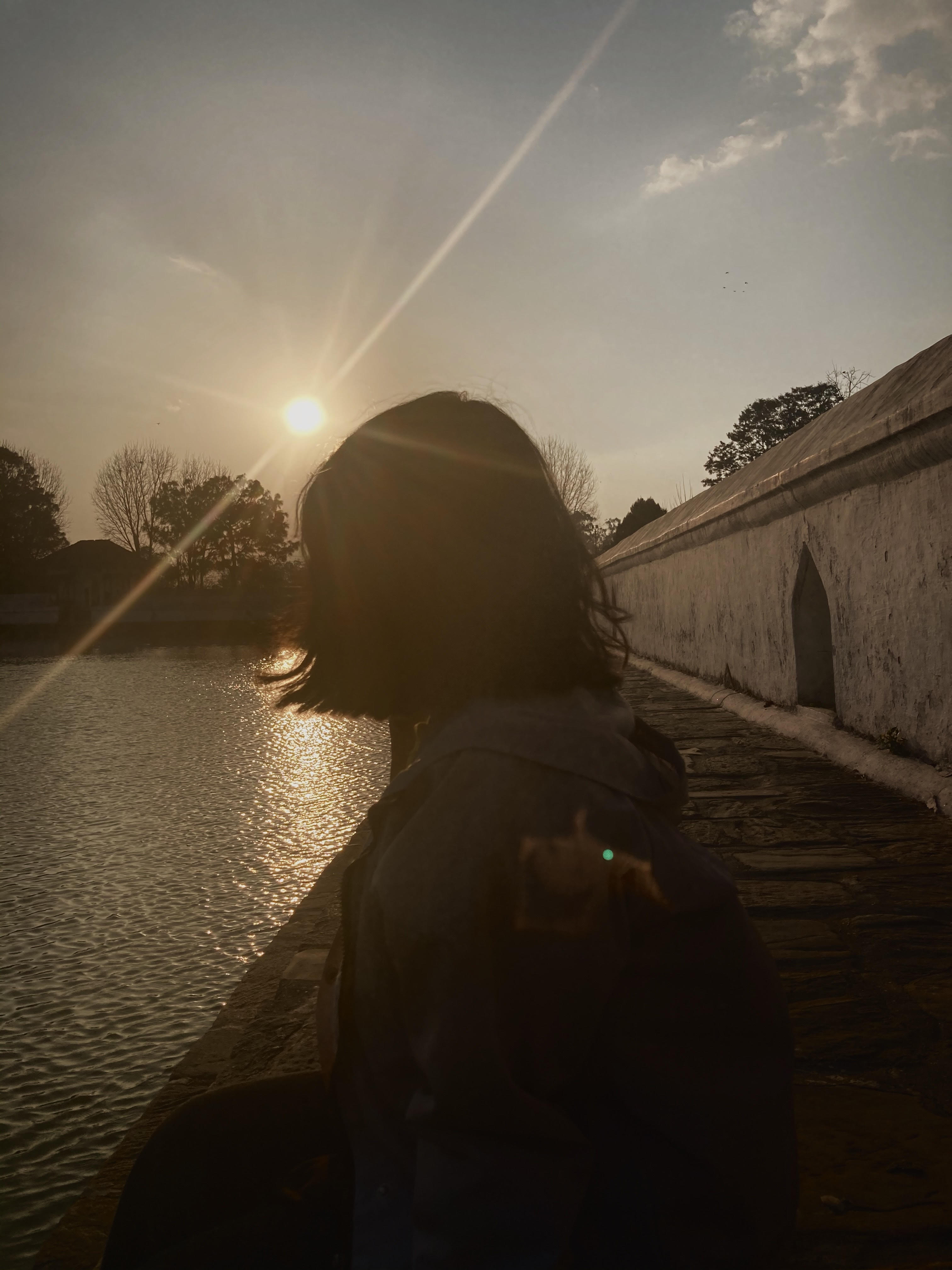 A silhouette of a person sitting by the water's edge during sunset. The sun is low on the horizon, casting a warm glow and creating a reflective shimmer on the water's surface.