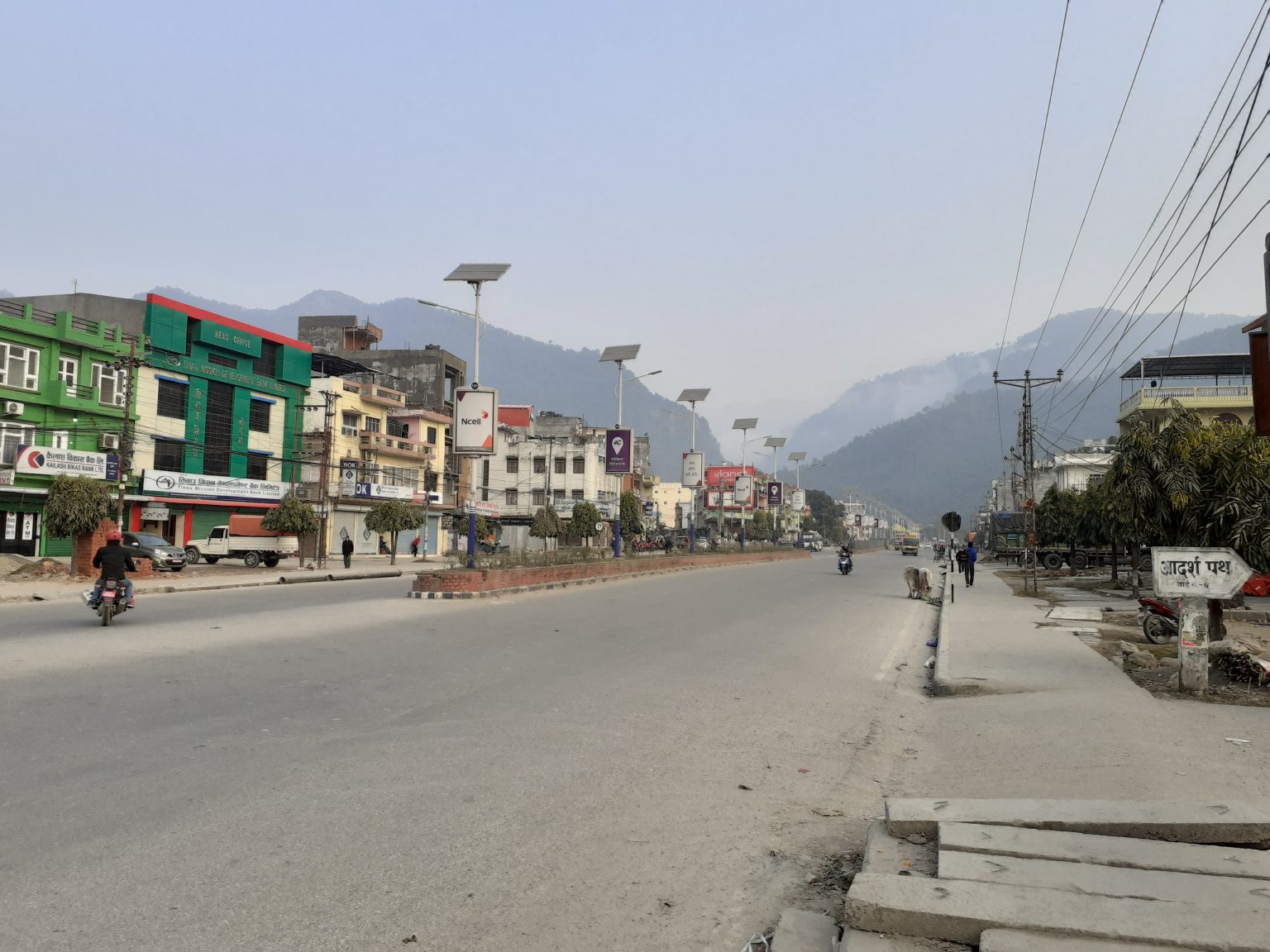 A quiet street scene in a mountainous area, featuring a wide road flanked by shops and buildings of various colors, including green, yellow, and gray.
