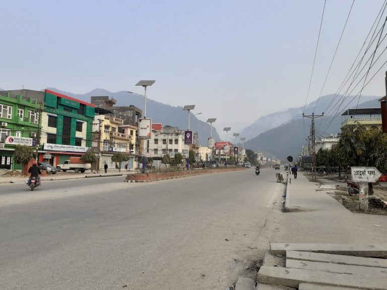 A quiet street scene in a mountainous area, featuring a wide road flanked by shops and buildings of various colors, including green, yellow, and gray.