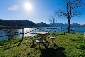 Picture of a table with wooden benches at the foot of an idyllic blue lake surrounded by large mountains on a sunny day.