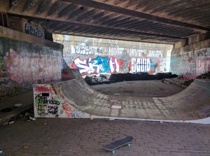 A DIY concrete skatepark with skateboard in foreground, a wooden mini ramp, under a graffiti tagged bridge in Worcester, Massachusetts.
