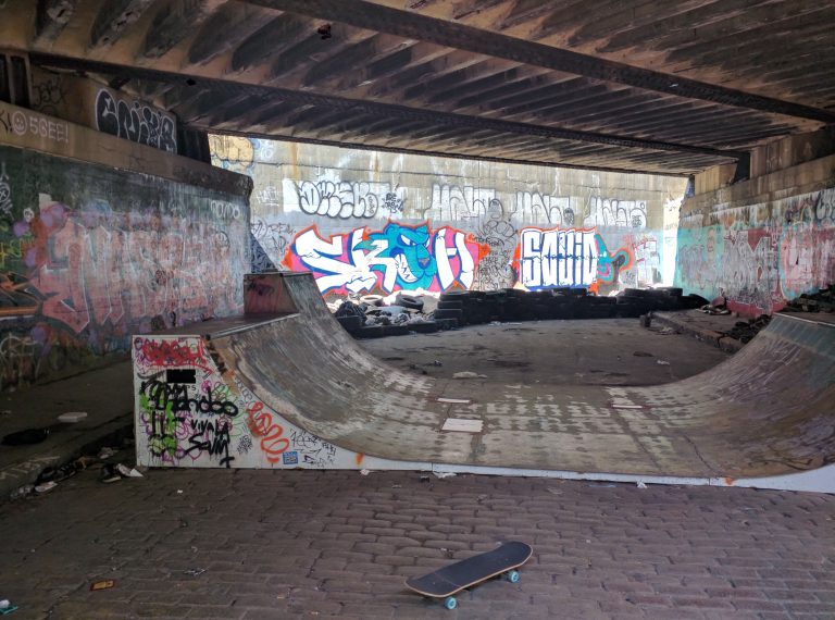 A DIY concrete skatepark with skateboard in foreground, a wooden mini ramp, under a graffiti tagged bridge in Worcester, Massachusetts.