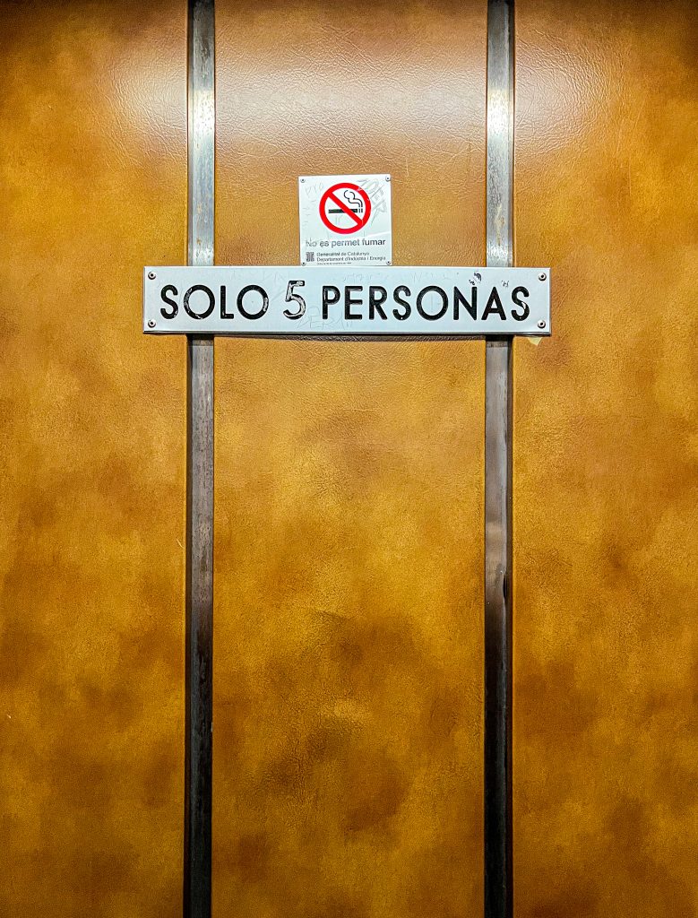 Inside a vintage elevator, a sign indicates, in Spanish, the maximum number of persons allowed in. Another sign indicates, in Catalan, smoking is forbiden.