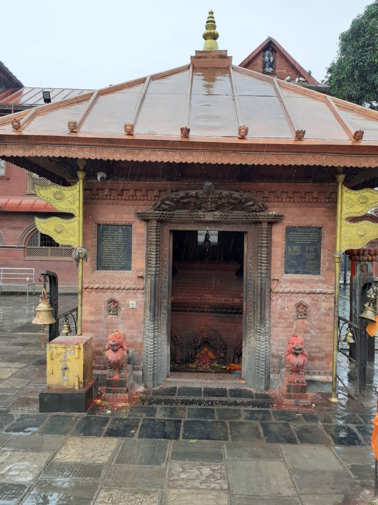 A small temple structure with a pitched roof and intricate carvings is situated in a rain-soaked environment. The temple features ornate decorations, including a golden finial on top and large stone lion statues at its entrance.