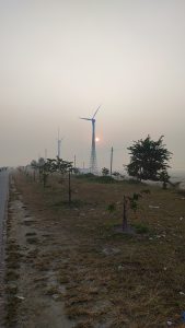 Windmill and Sunrise,  The sun is just off the horizon in a foggy sky.  We’re standing on the edge of a road, looking down the road to the horizon. Four giant industrial windmills are some distance from the road.
