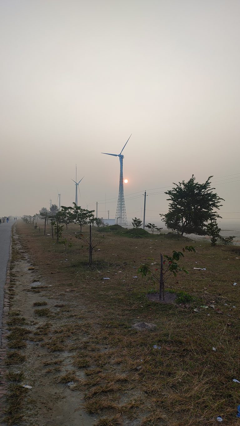 Windmill and Sunrise,? The sun is just off the horizon in a foggy sky.? We’re standing on the edge of a road, looking down the road to the horizon. Four giant industrial windmills are some distance from the road.