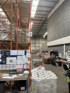 Warehousing environment storing, picking and packaging. Towering shelves on the left, brick wall on the right. Lots of industrial desk space.