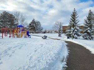 Winter snow along with beautiful nature. Snowy school yard, sidewalk on right curving across to the left behind some playground equipment.
