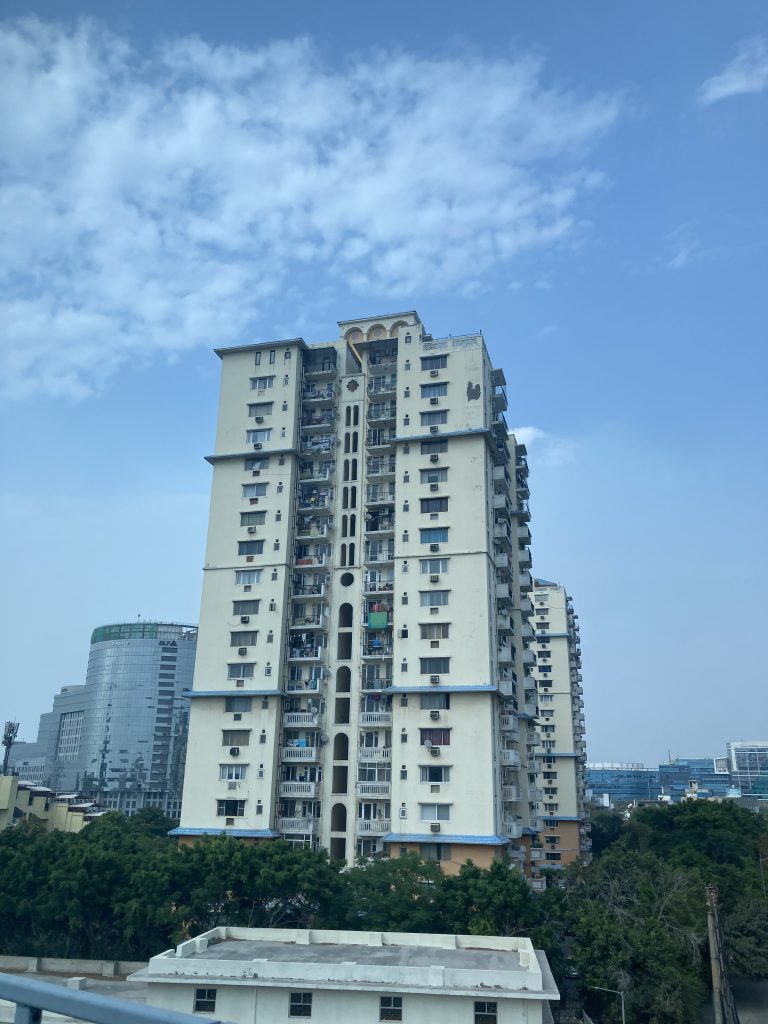 City Buildings, highrise apartment building that’s almost the same color as the sky. Glass skyscrapers in the background.