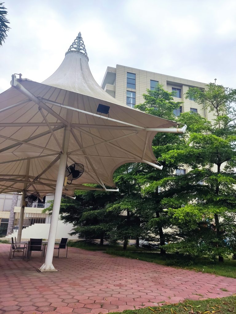 Camera near the ground looking up at an industrial umbrella with speakers and fans inside. It’s on a brick patio. There are trees on the right, and a highrise building in the background.