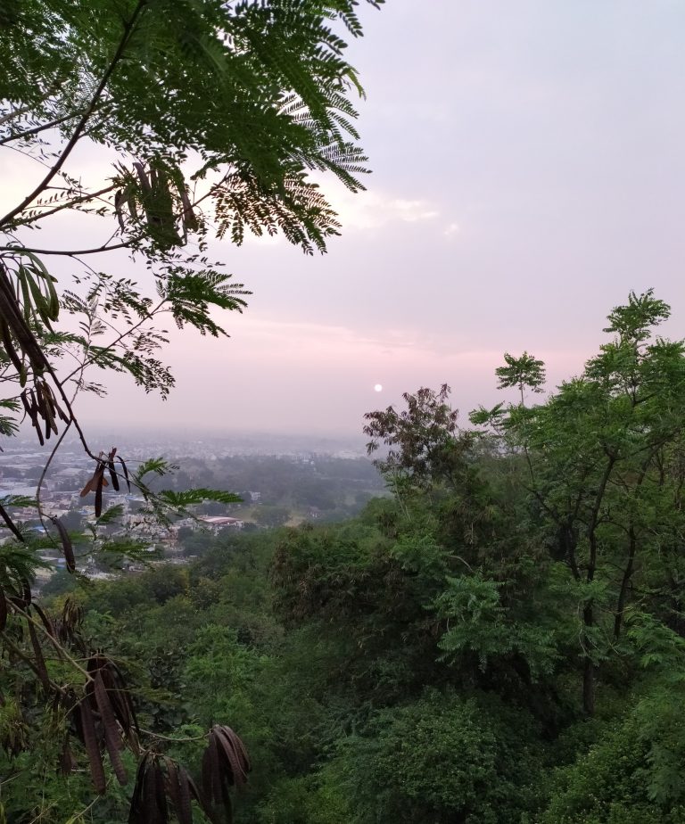 Long view from a hilltop. Small hill in foreground, but then in the distance a village.? The sun is small and just off the horizon. Cloudy sky.