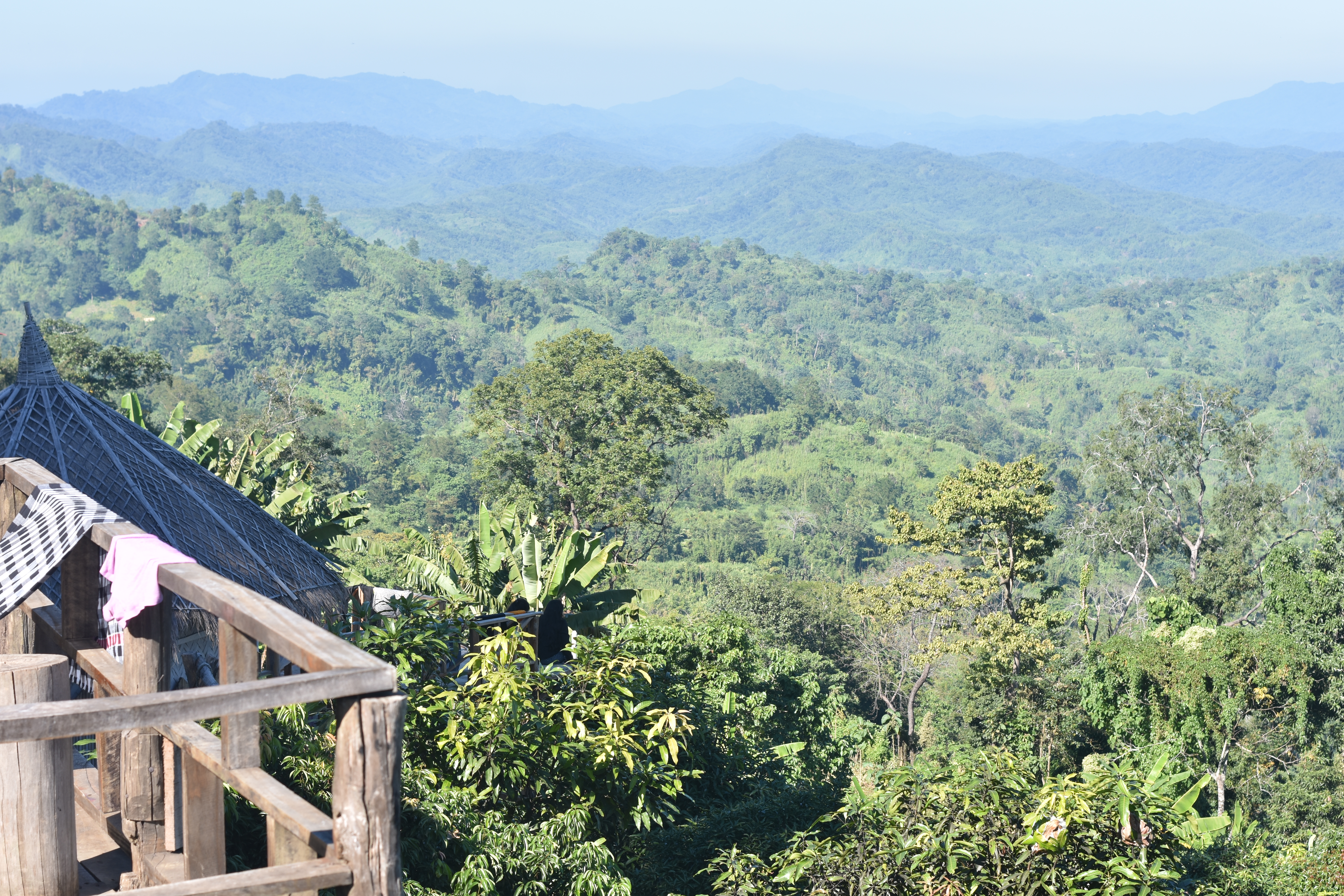 At Sajek, Bangladesh, a long distance view over the forest to rolling hills. In the left side of the frame we can see a bit of deck, like someone might stand on to observe.