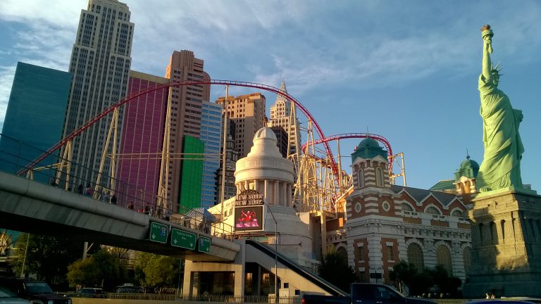 Las Vegas strip. Looking up at colored high rise buildings, the statue of Liberty on the right, colored pipes from a roller coaster weaving throughout.