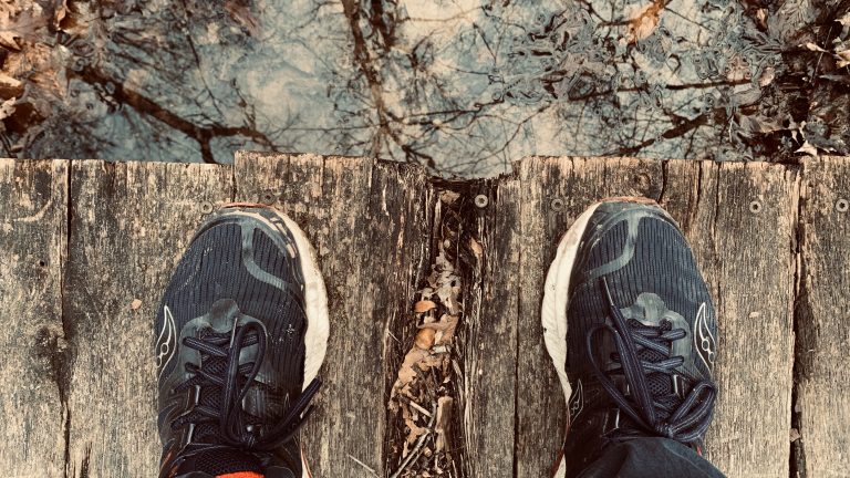 Running Shoes on Bridge, New River Gorge National Park, Fayette County, West Virginia. Camera is near the knees, looking down at the shoes.? The toes are about an inch from the edge of the bridge. The bridge is made of very old wood. There’s water down below, reflecting the sky.