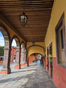 View larger photo: A covered walkway in San Miguel Allende. Looking the long way down the walkway, arches on the left opening to an open space, wall on the right with doors.