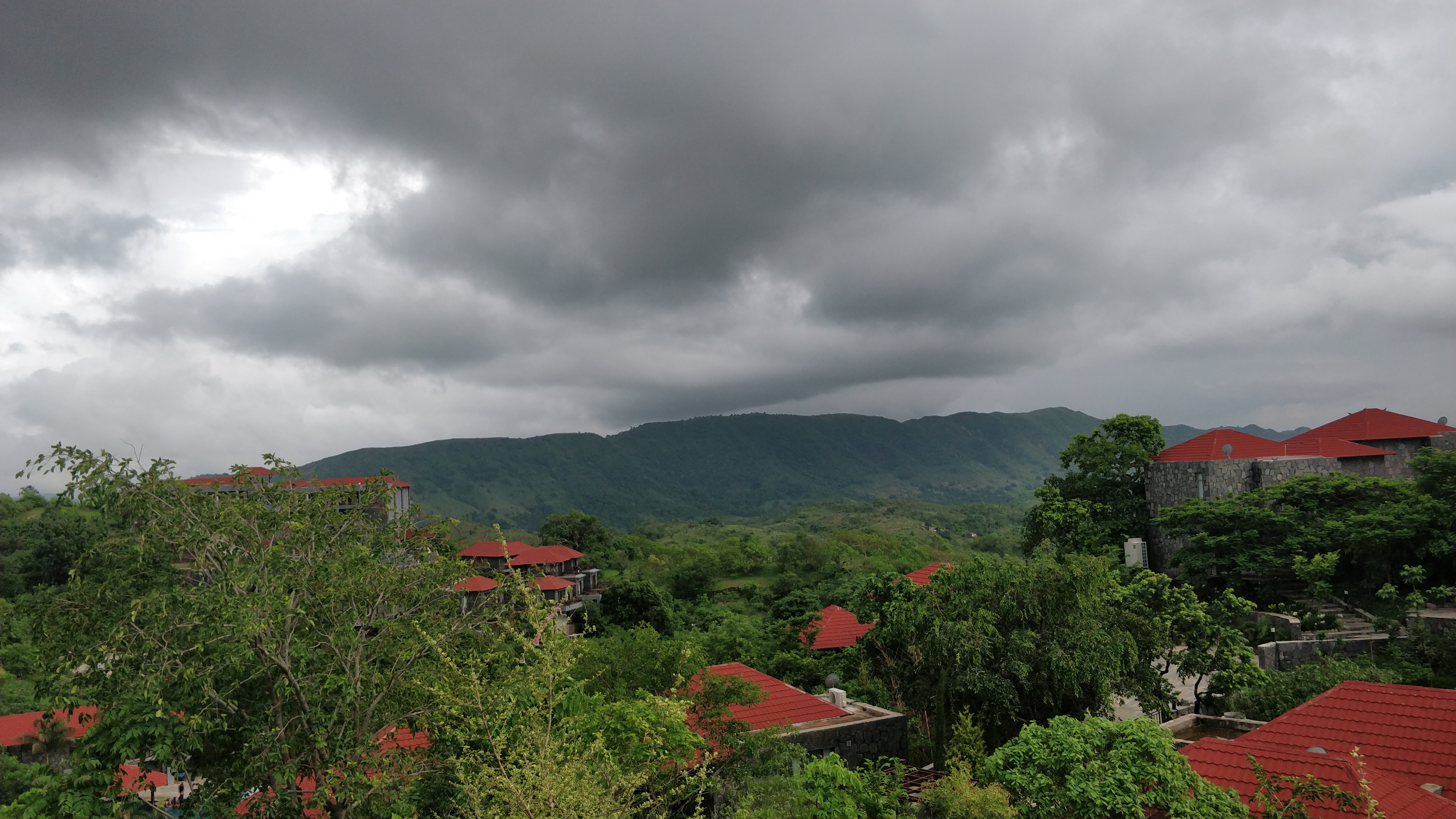 A panoramic view of a mountainous landscape under a cloudy sky. Dark gray clouds dominate the sky, with hints of light peeking through. In the foreground, lush greenery surrounds a cluster of buildings with red roofs nestled among the trees, while rolling hills stretch into the distance, creating a serene, rural atmosphere.