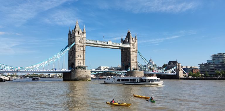 Tower Bridge, London. Photographer is on the water, it’s a sunny day, and in the foreground is a large tour boat and two single-person kayaks.