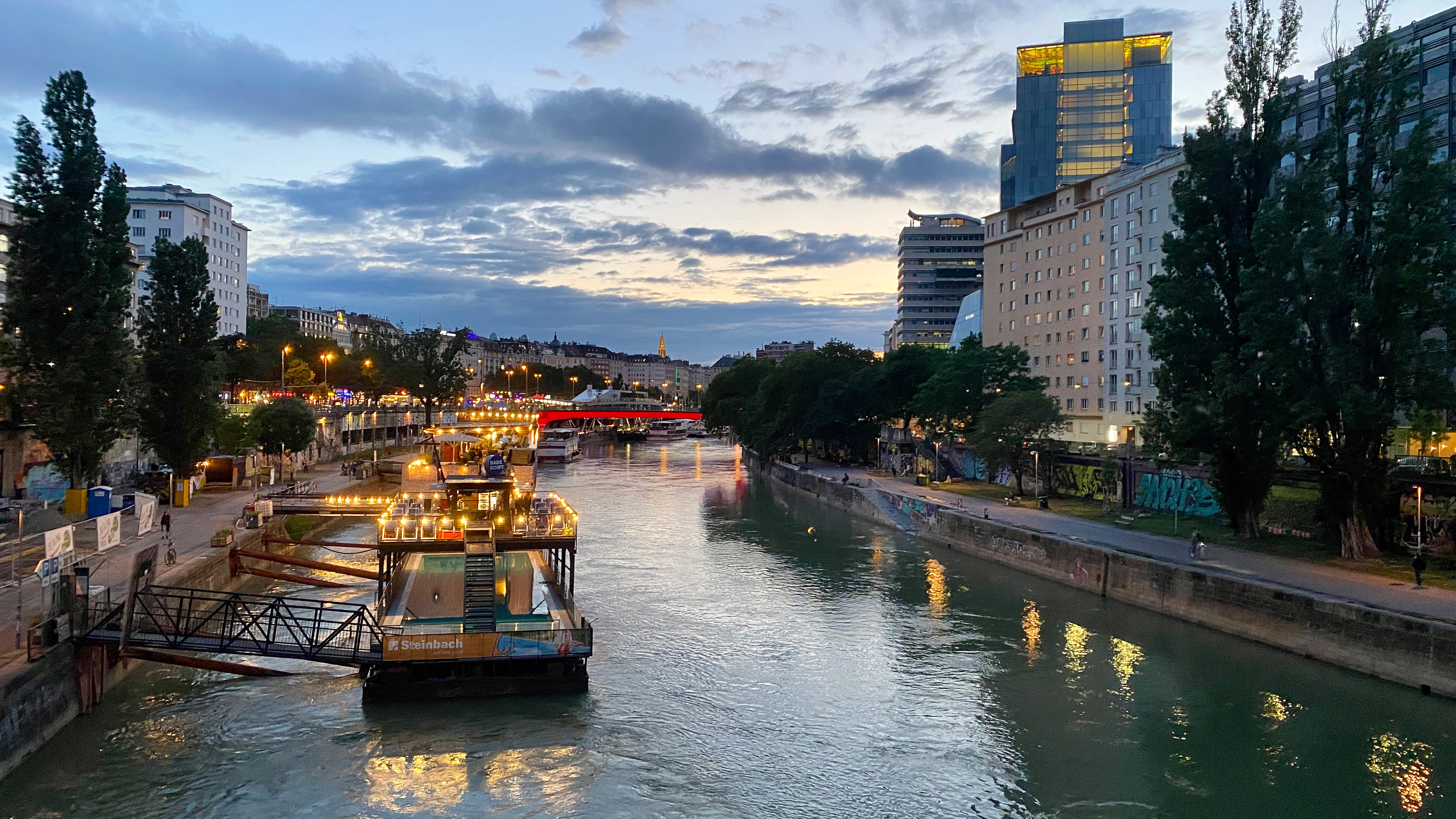 Vienna, view from a bridge in the dawn. Looking down the river, boats along the shore with lights, like party boats. Hotels and tall buildings along the right.? Looking toward the dawn.