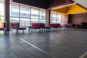 The hallway features red sofas and tables placed by the windows, creating a cozy and inviting atmosphere. The wide hall has a yellow element on the ceiling that adds a pop of color to the space. The lighting is bright and natural, highlighting the details of the furniture and the architecture of the building.