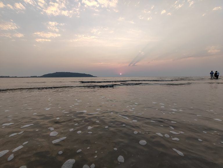 Sunset at the beach. The camera is very close to the water and the sun looks very far away, just a dot. There’s an island on the horizon, and a few people standing in the water about knee deep.