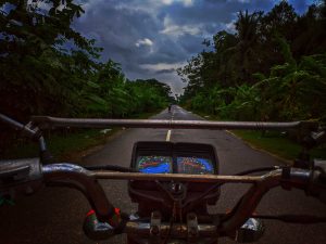 Camera view is sitting on the gas tank of a motorcycle, looking ahead over the handlebars on a straight road, with a cloudy sky after the rain overhead. The wet asphalt glistens, reflecting the dim sunlight, creating a serene and atmospheric scene. The road is narrow with jungle on each side.
