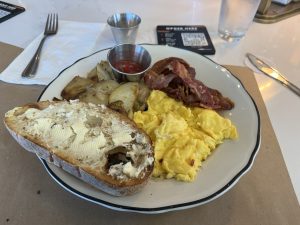 Breakfast plate. Bespoke sourdough bread with butter, closest to the camera. Just to the right scrambled eggs, at about 1 o'clock a pile of bacon, then at 12 o'clock a little metal tub ot catsup, and then at 9 o'clock fried potatoes.