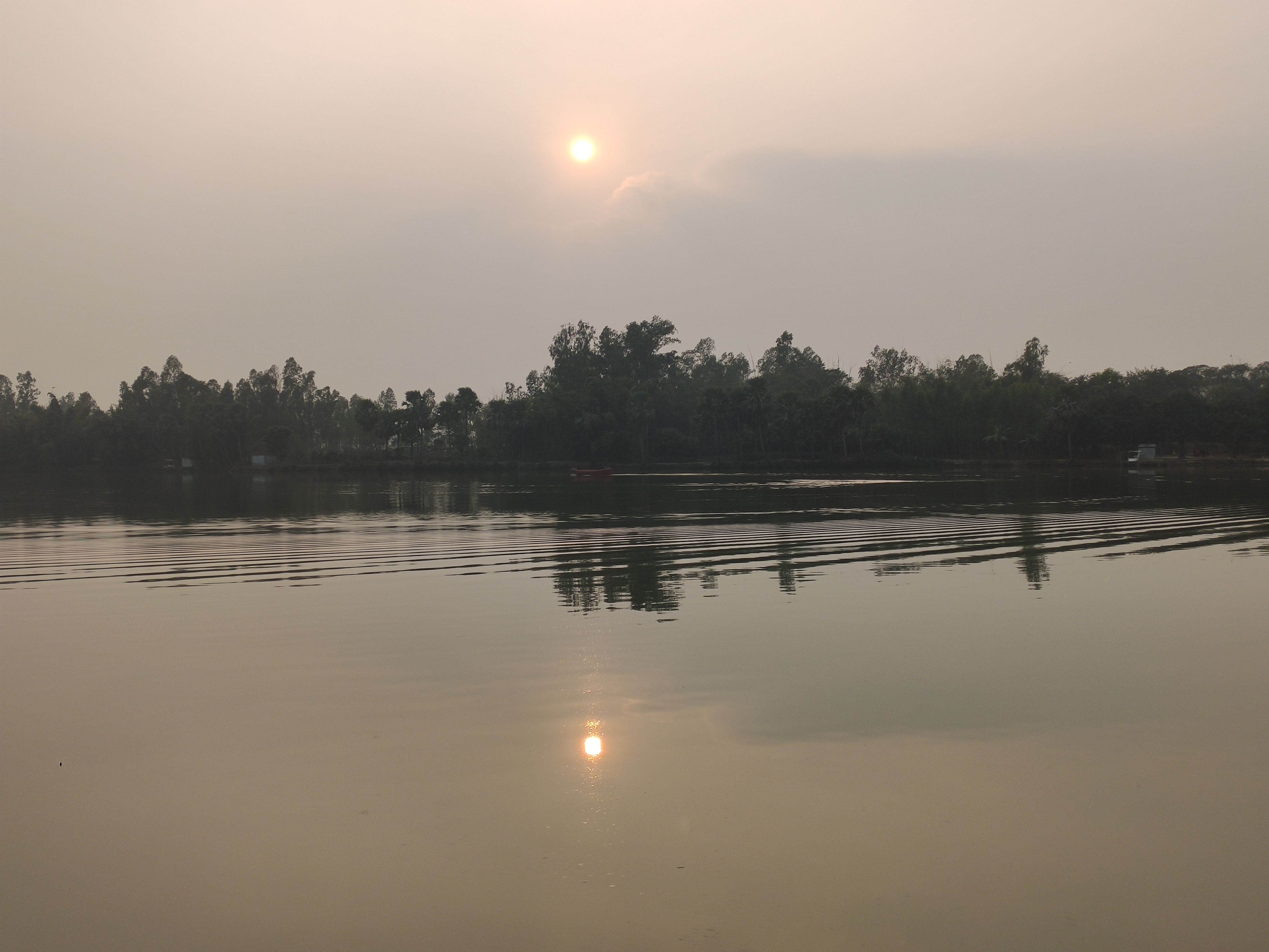 A calm lake or river in the foreground, with forest on the far side. The sun is just over the horizon but mostly hidden behind clouds so the world is dim.