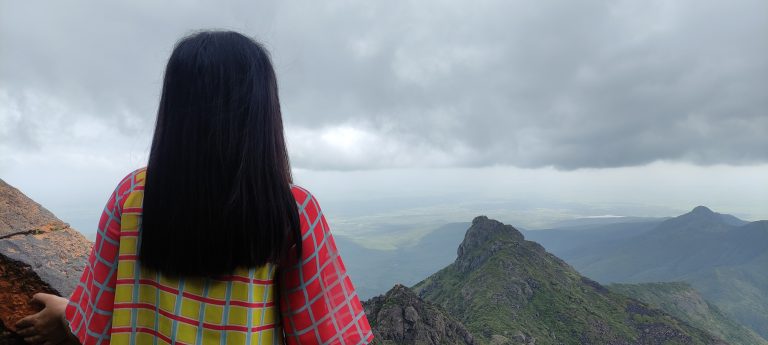 Woman standing by a wall, looking over a vast expanse of mountains, stormy sky above