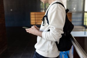 View larger photo: A man in a white shirt and black backpack leans against a chrome railing inside a building while looking at his phone. The man appears relaxed, with his weight resting on one leg and his other foot crossed over it. He holds the phone in one hand and seems to be scrolling through its contents.