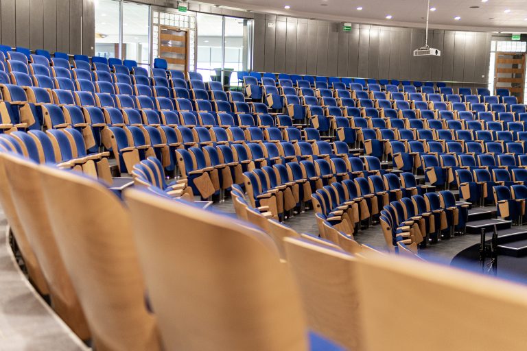 An empty auditorium in a university is furnished with wooden chairs that have blue cushions. The chairs are neatly arranged in rows, facing a stage or a podium that is not visible in the frame. The room is empty of people, and the lighting is relatively bright, daytime or artificial lighting. The overall atmosphere of the scene is academy