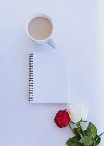 Taza de café con libreta y rosas a un costado. Todo sobre fondo blanco. Flatlay.