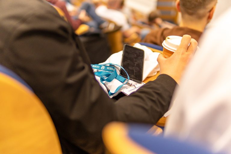 Blue lanyard with WordPress logo. Next to it stands an orange takeaway coffee mug. The photo is taken in an auditorium during a conference or lecture.