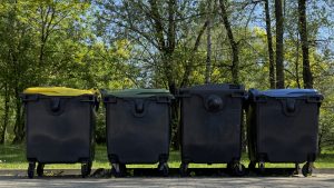 View larger photo: Four waste garbage cans standing in a row against a background of greenery and trees. Eco-friendly trash bins.