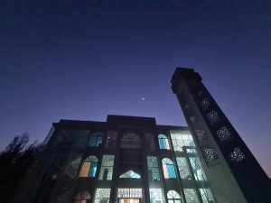 Looking up at the night sky, a tiny crescent moon visible.  In the foreground is a tower and a three storey building lit from the inside with each window having a unique geometric shadow pattern on it.
