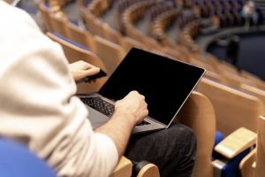 View larger photo: In an empty auditorium, a man sits with a laptop on his lap. The wooden chairs around him have blue cushions, adding a pop of color to the otherwise plain setting. The man's face is not visible in the frame. The lighting is relatively dim, with some natural light coming in from the windows behind the man. The overall mood of the scene is calm.