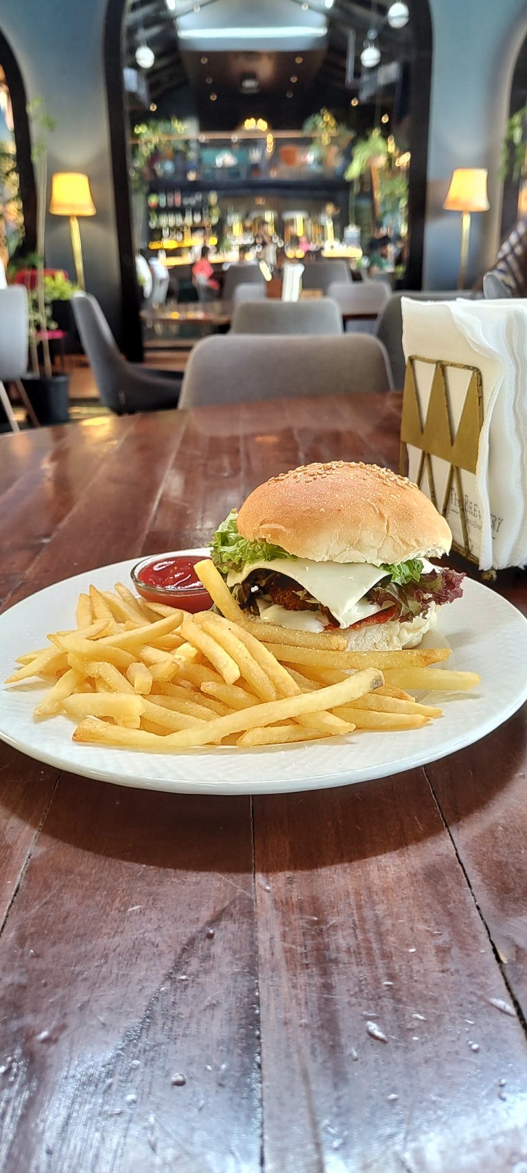 On a wooden table in a restaurant, a plate with french fries, a small bowl of catsup. amd a burger with lettuce, cheese, tomato, and a sesame seed bun.